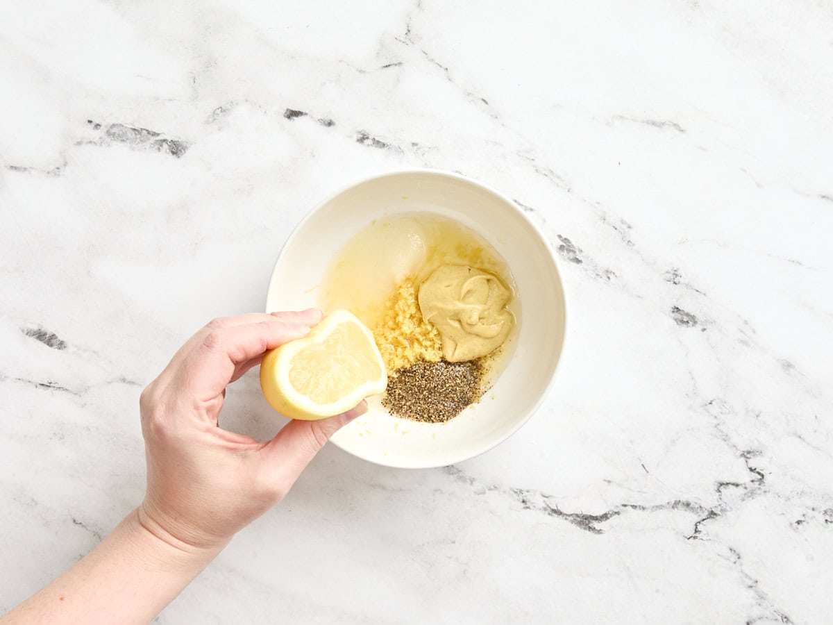 Eine Hand drückt eine halbe Zitrone in eine Schüssel Apfelessig, Senf, Zucker und Gewürze.