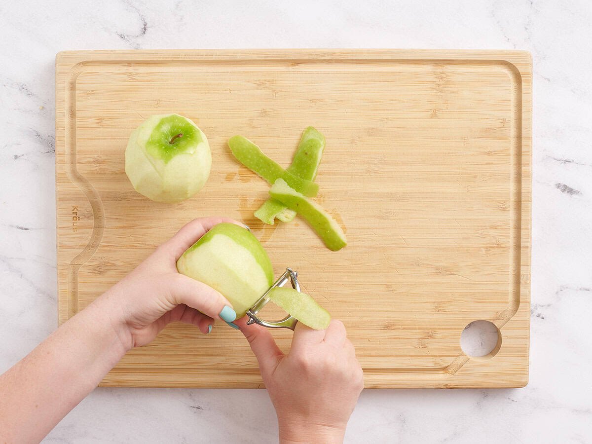 Eine Hand mit einem Gemüseschäler, um einen grünen Apfel zu schälen.