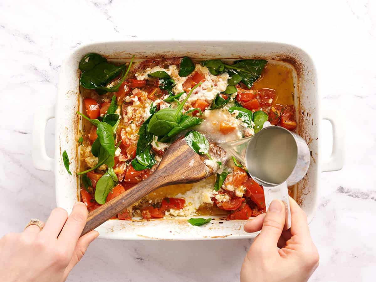 Eine Hand gießt zurückgebliebenes Nudelwasser in eine Auflaufform mit gebackenen Tomaten und Feta-Käse.