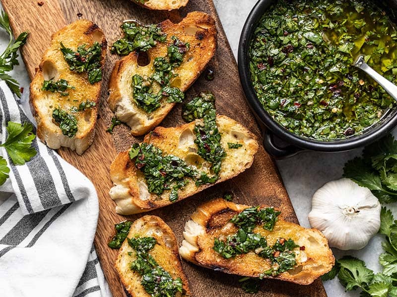 In Scheiben geschnittenes, geröstetes französisches Brot auf einem Schneidebrett, beträufelt mit Chimichurri und einer Schüssel Chimichurri-Sauce als Beilage.
