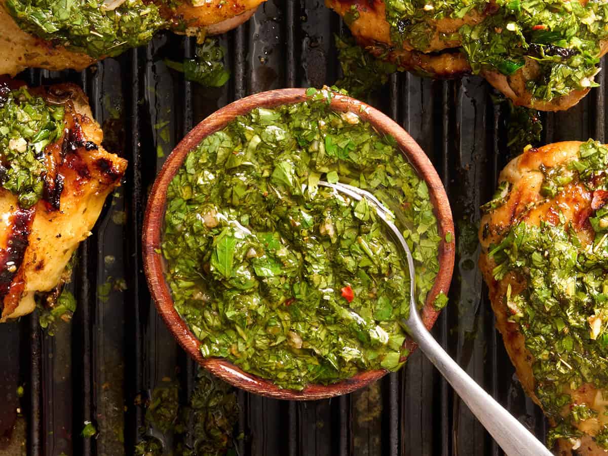Blick von oben auf eine Schüssel Chimichurri auf einem Grill mit einem Löffel und gegrillten Hähnchenschenkeln.