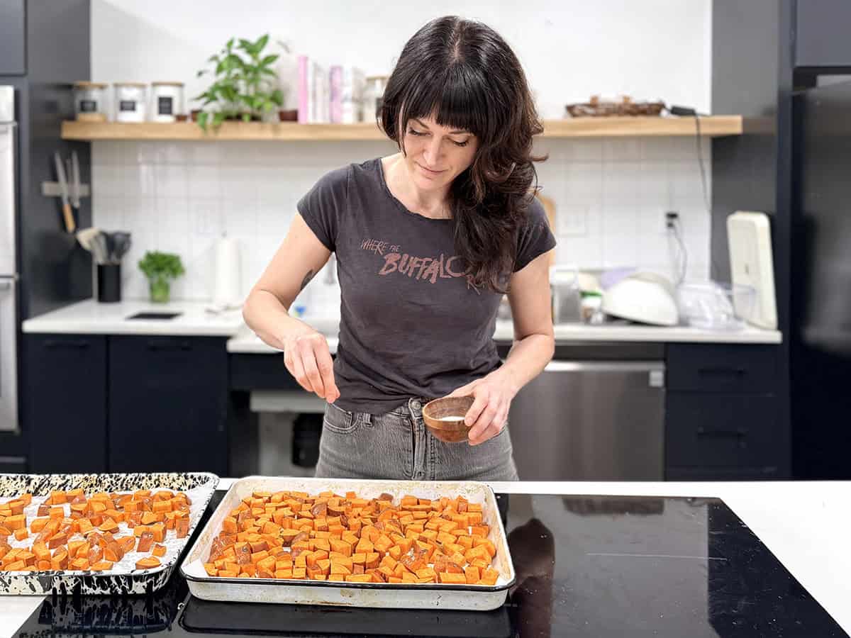 Eine Frau streut Salz über ein Blech mit gewürfelten Süßkartoffeln in einer Küche.